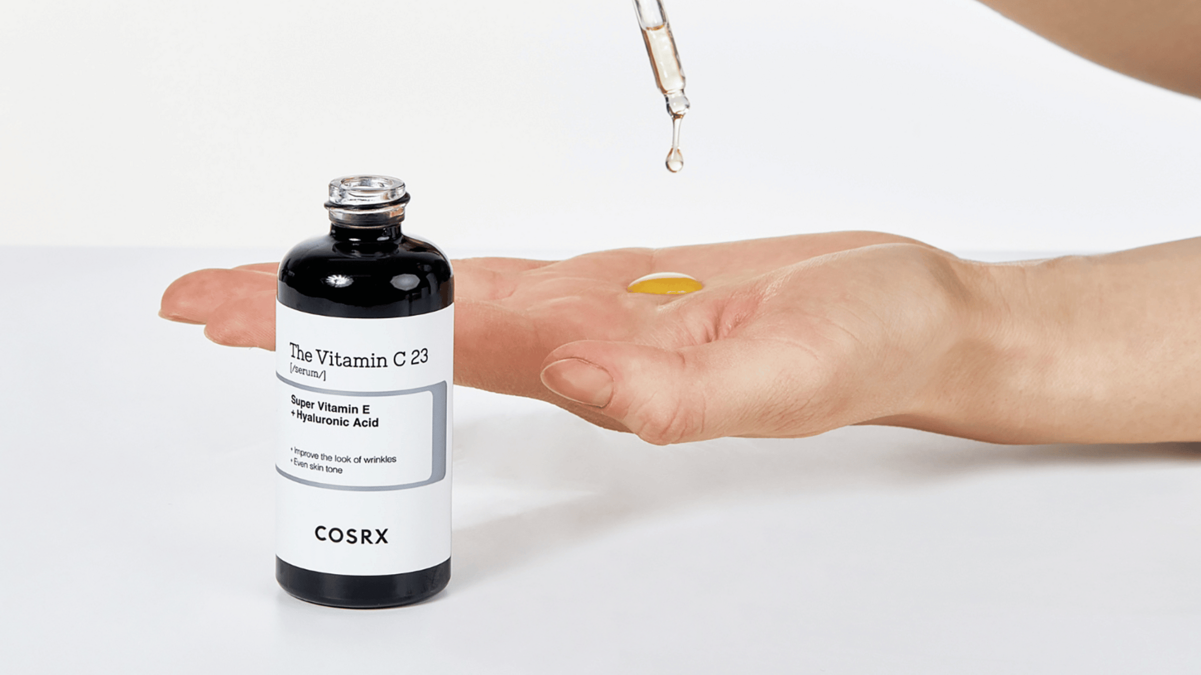 COSRX The Vitamin C 23 Serum med Super Vitamin E och hyaluronsyra. En hand håller serumet, och en pipett droppar gul vätska i handflatan. Produkten är designad för att reducera rynkor och jämna ut hudtonen, perfekt för en strålande hud.