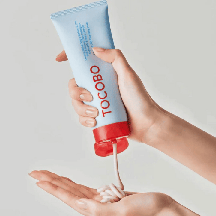 En person håller i en tub av TOCOBO Coconut Clay Cleansing Foam. Tuben är ljusblå med en röd kork och texten "TOCOBO" på framsidan. Produkten trycks ut ur tuben och formas som ett schampo i handen. Bakgrunden är neutral och ger fokus på produkten.