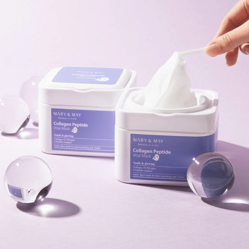 En hand tar upp en mask från en burk med MARY & MAY Collagen Peptide Vital Mask. Masken är avsedd för daglig användning för att ge näring och glöd åt huden, med kollagen och peptider för att stödja hudens ungdomlighet.