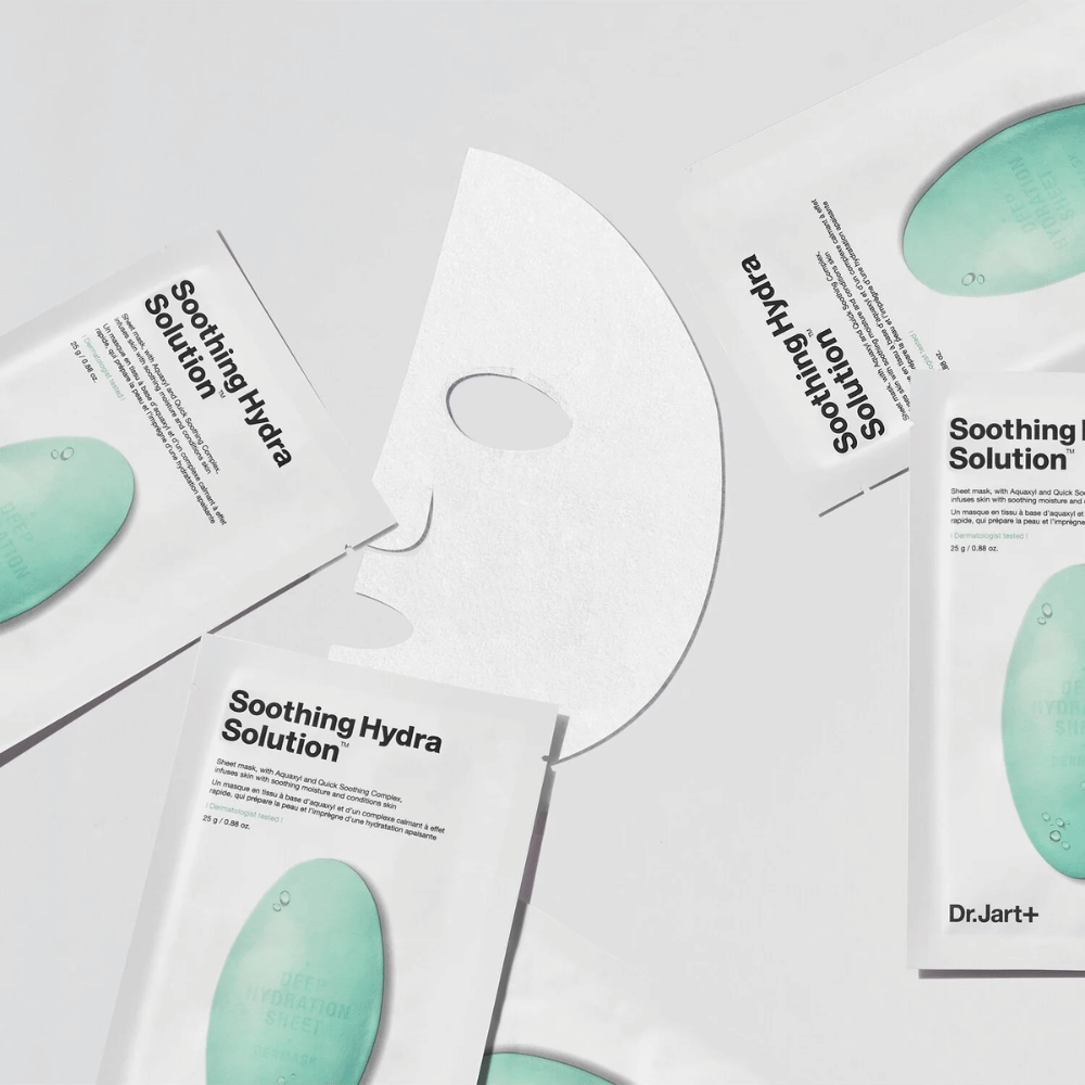 Flera förpackningar av "Soothing Hydra Solution" ansiktsmasker från Dr.Jart+ är utspridda på en ljus yta. En utvecklad mask är placerad i mitten, vilket visar dess vita och hålade struktur.