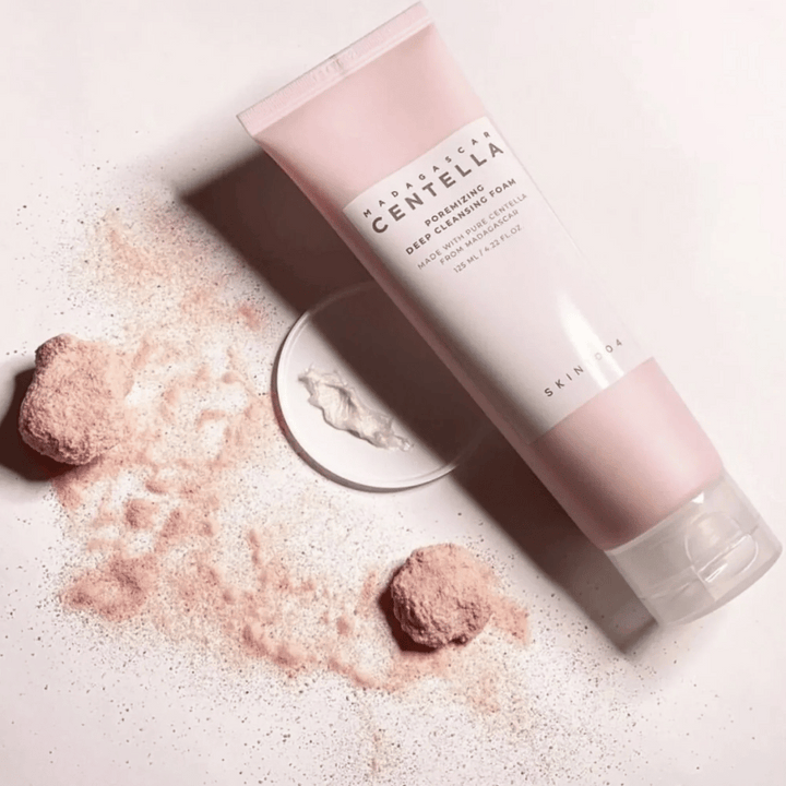 Madagascar Centella Poremizing Deep Cleansing Foam presenteras i en rosa tub. Tuber ligger på ett ljust underlag tillsammans med rosa pulver som ger en känsla av renhet och fräschör. Produkten är designad för att djup rengöra huden med hjälp av ren centella från Madagaskar.