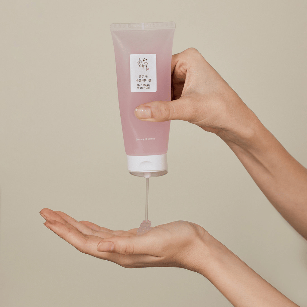 Bilden visar en person som håller en tub av "Red Bean Water Gel" från Beauty of Joseon och klämmer ut en genomskinlig gel i sin hand. Produkten ser ut att vara avsedd för hudvård och har en attraktiv, mjuk rosa förpackning.